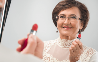 Older woman looks in the mirror, holding her lipstick and smiling because her FOY dentures allow her lipstick to look amazing